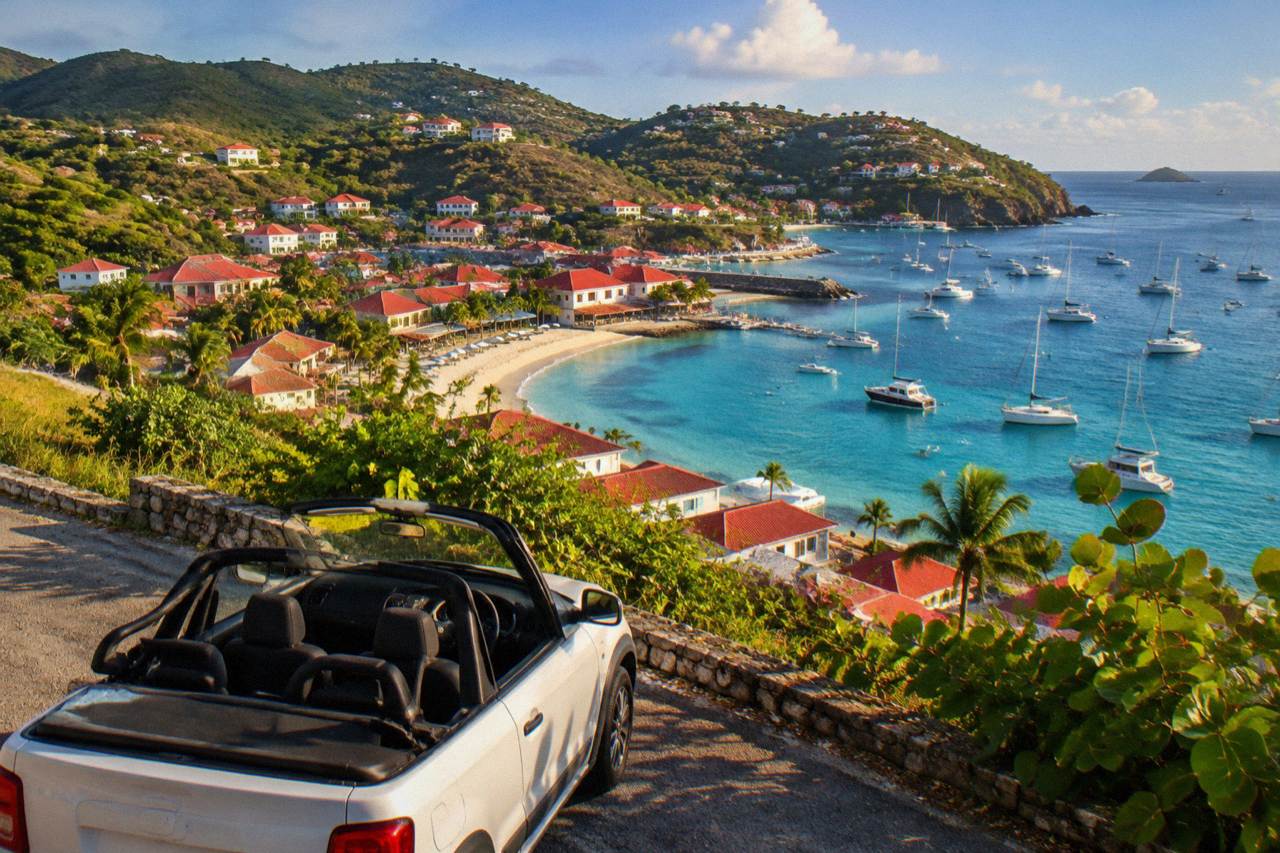 A small open-top rental car parked on a narrow hillside road above a Caribbean bay with red rooftops below