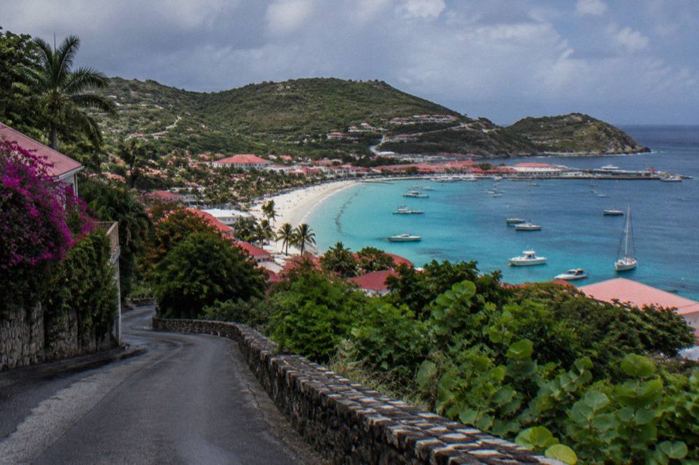 A steep narrow road curving down through green hills toward a small Caribbean harbour town