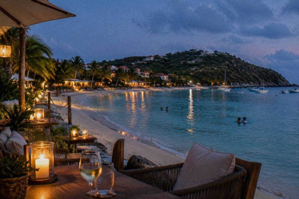 A beach club terrace at dusk on Saint-Jean bay, St Barts, with lights reflecting on calm water