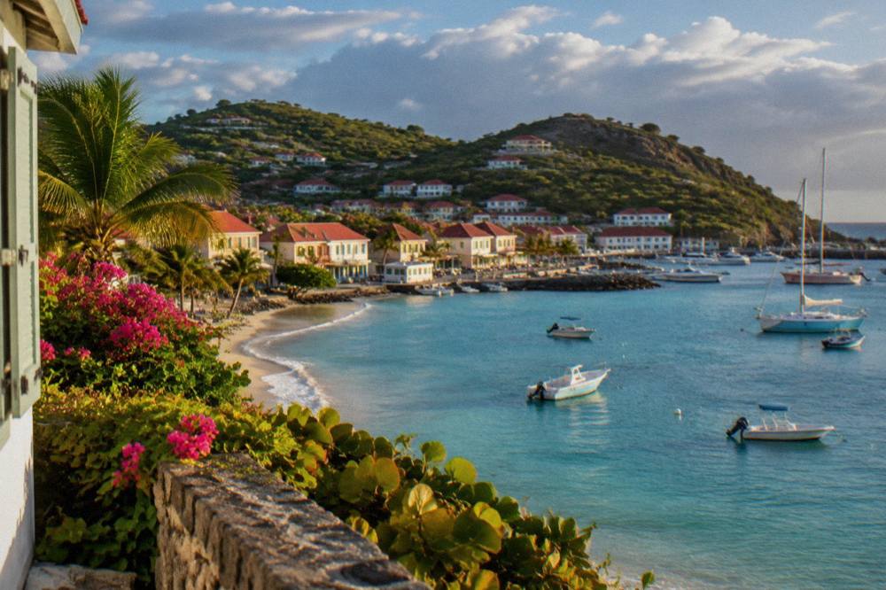 A green hillside descending to a curved sandy beach with calm turquoise shallows and a few anchored boats