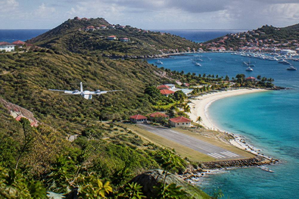 A small turboprop aircraft on a short runway beside a curved Caribbean beach between green hills