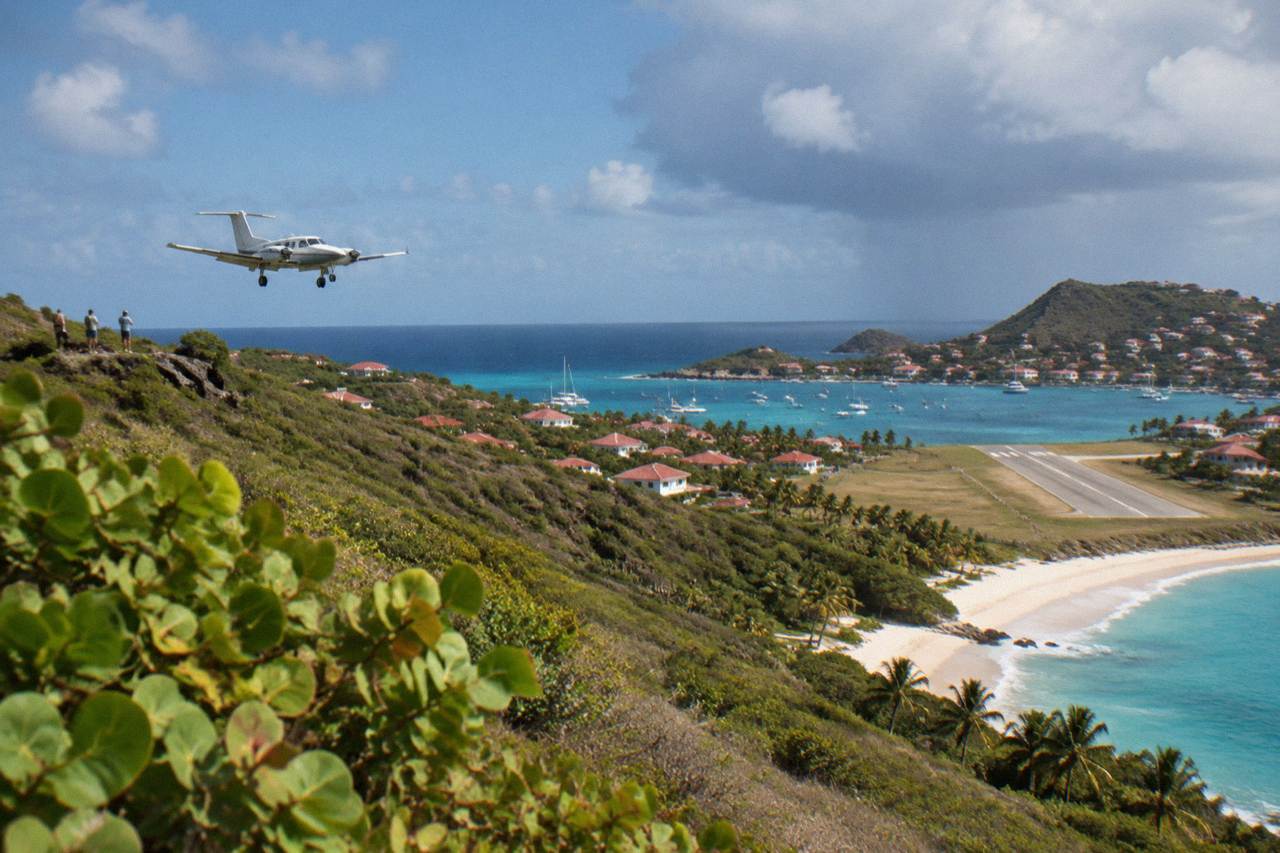 A light aircraft flying low over green hills toward a small Caribbean island airstrip beside turquoise water