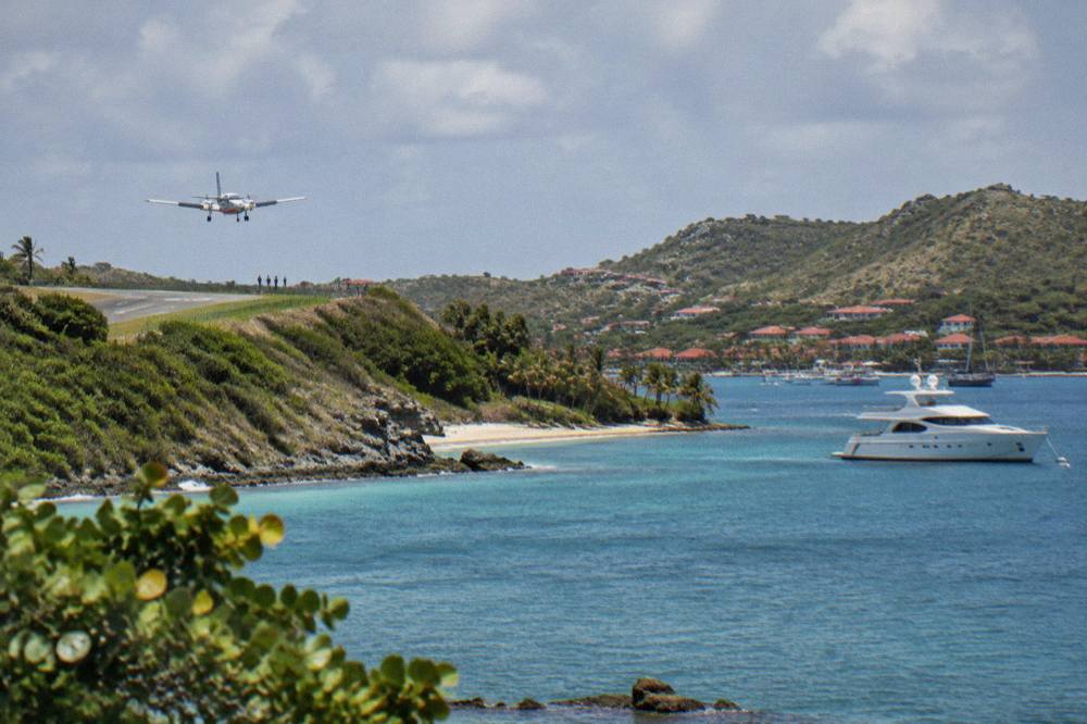 A white motor yacht anchored in a calm Caribbean harbour with hills and red rooftops behind it