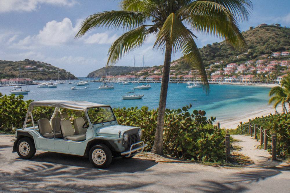 An open-air Mini Moke parked beside a palm tree near a beach entrance on St Barts