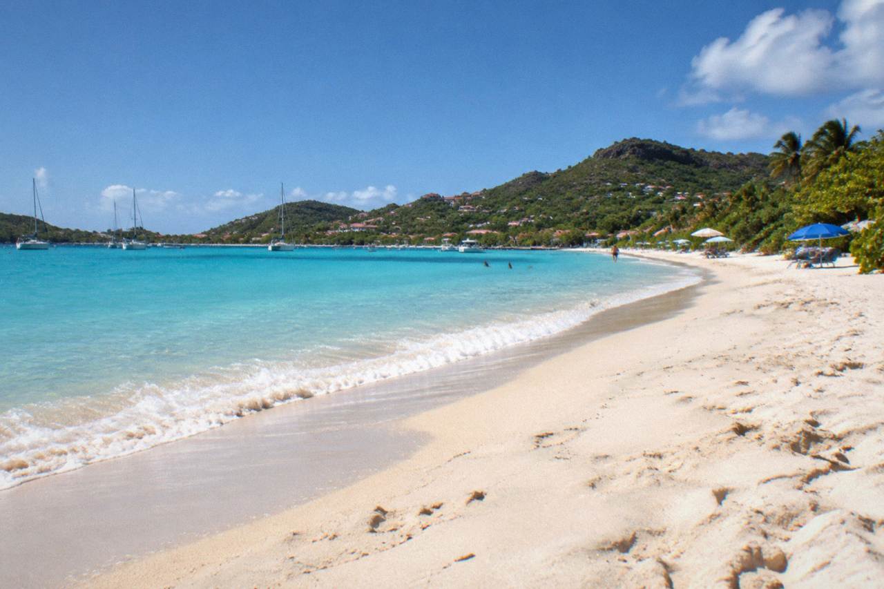 A wide sandy beach backed by low green hills under a clear sky, with a few scattered beach umbrellas near the waterline
