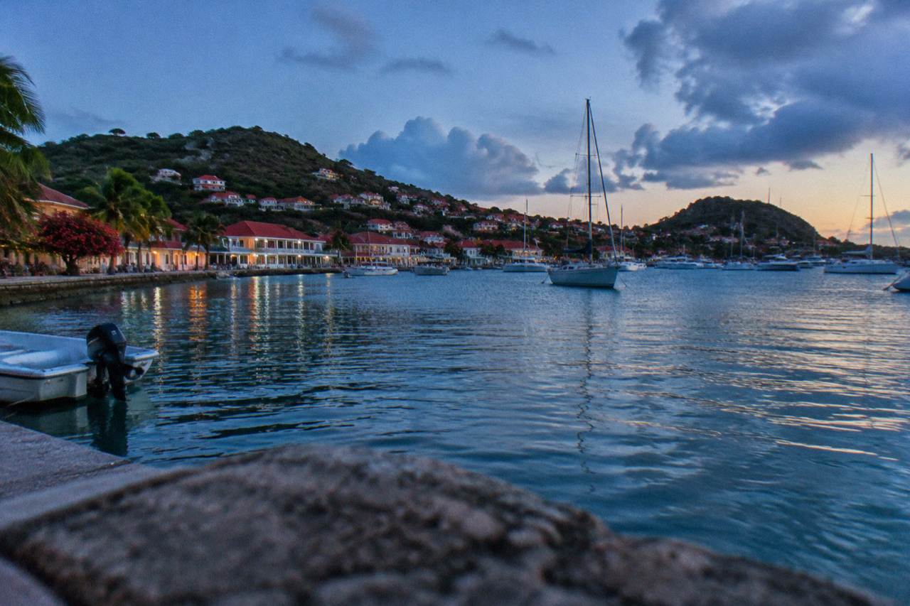 A red-roofed Caribbean harbour town with hills rising behind a bay of moored sailboats