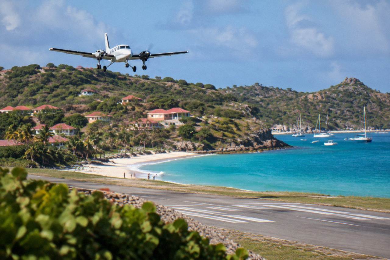 A small turboprop aircraft on a short runway beside a Caribbean beach with green hills behind it