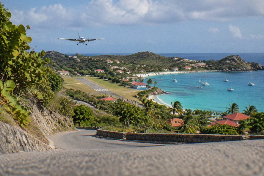 A steep green hillside road descending toward a small airstrip and a turquoise bay in the Caribbean