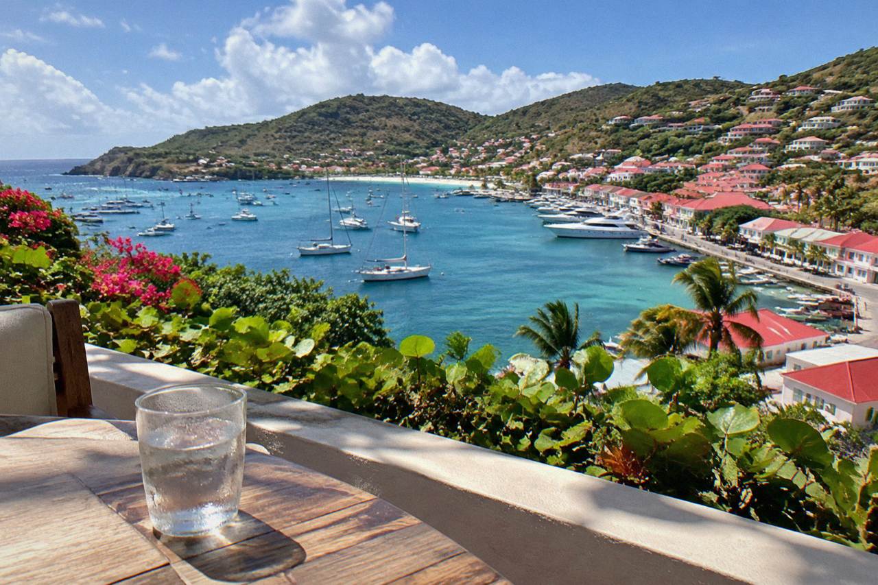 A glass of water on a sunny terrace overlooking a Caribbean harbor with hills and boats