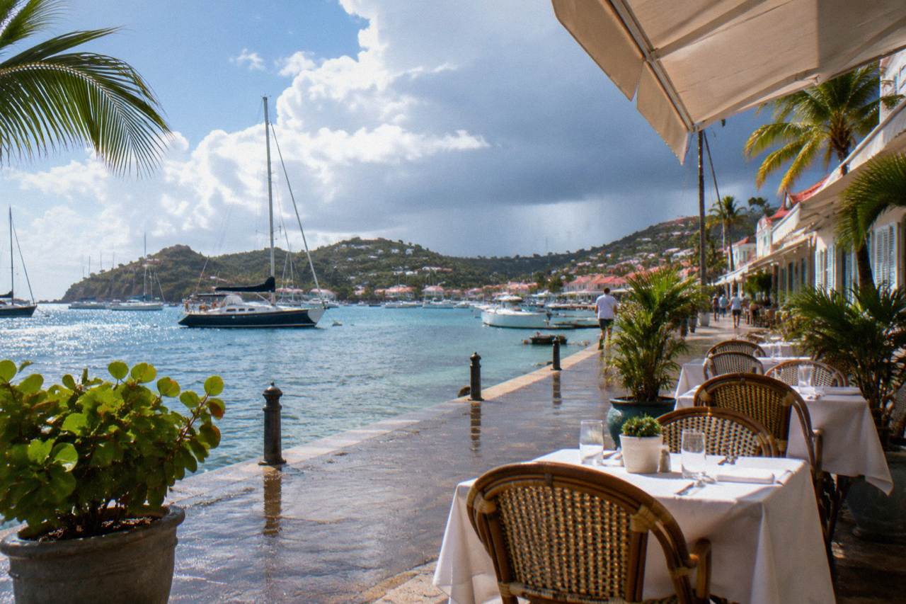 A harborfront terrace with white tablecloths and rattan chairs looking out over sailboats in a small Caribbean port