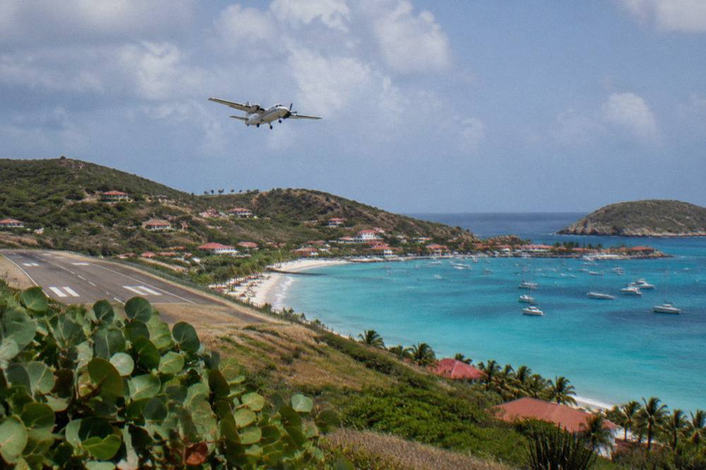 A turboprop climbing away from a Caribbean island airport with turquoise water along the shoreline