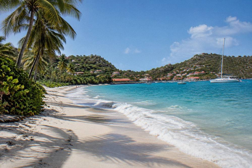 A yacht anchored in a small Caribbean harbour with hills and red roofs behind it