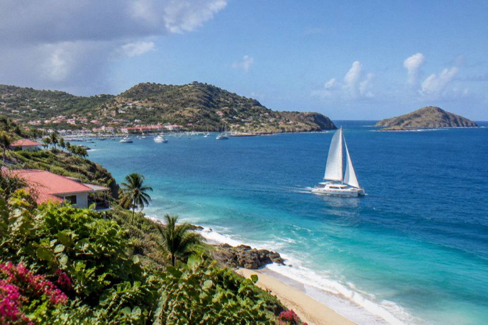 A catamaran under sail on open blue water between two Caribbean islands