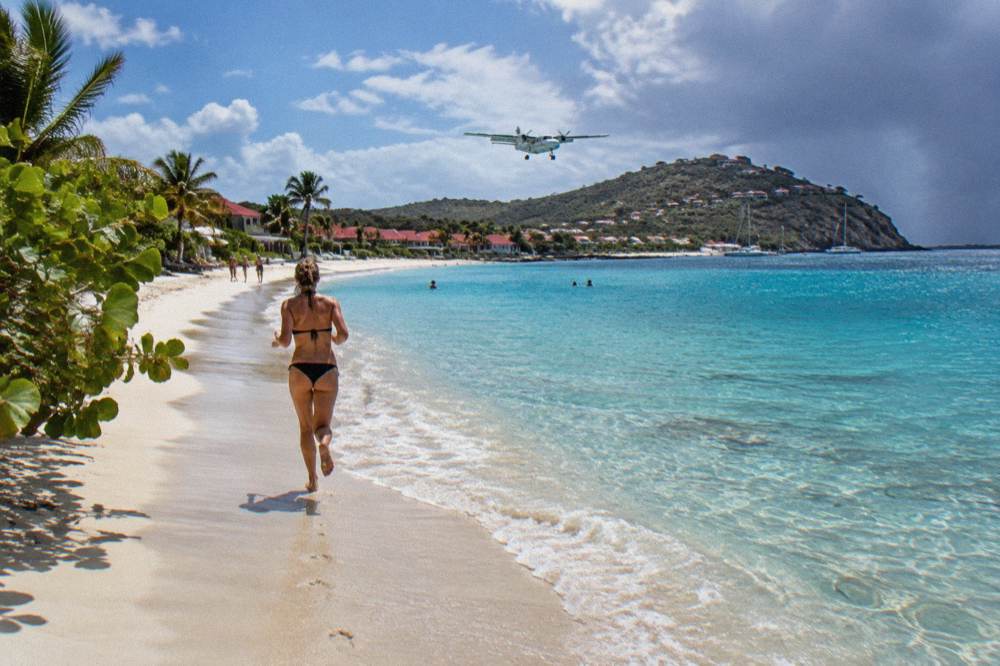 A small turboprop aircraft landing over the beach at St Jean with hills behind