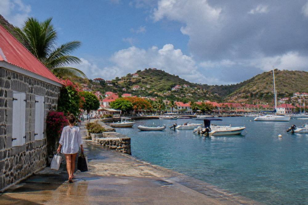 A shopper carrying bags past a stone building with a red roof in Gustavia