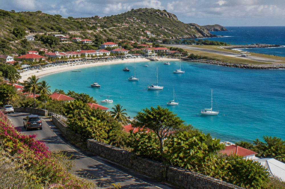A hillside road overlooking a bay with a small aircraft on a runway beside the water