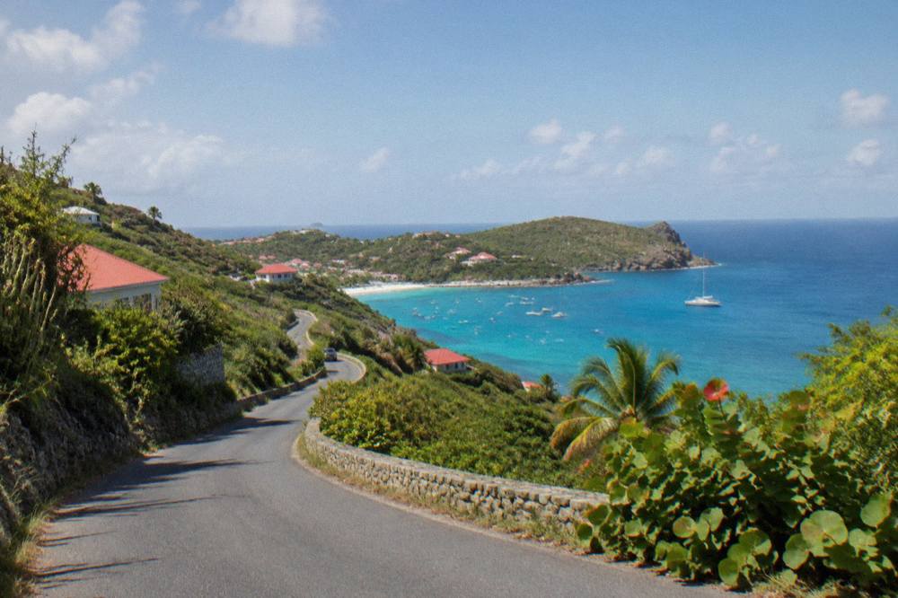 A narrow paved road curving between green hills with a glimpse of sea beyond