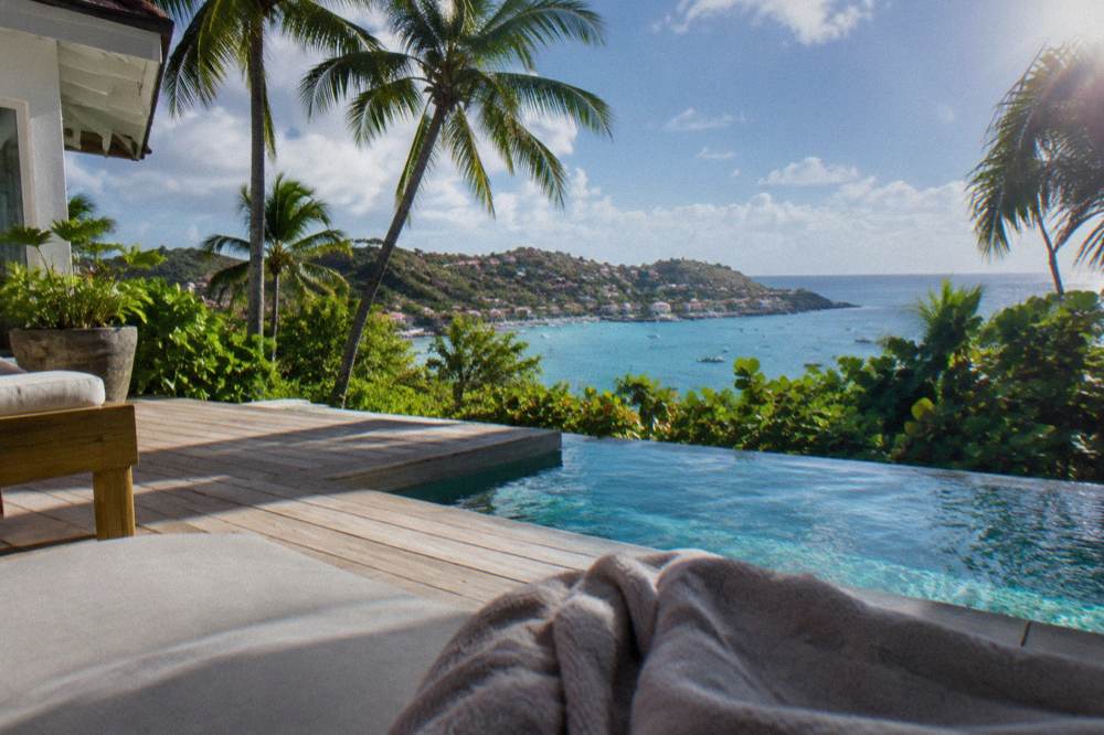 A white-walled villa with a wooden deck and pool overlooking the sea through palms