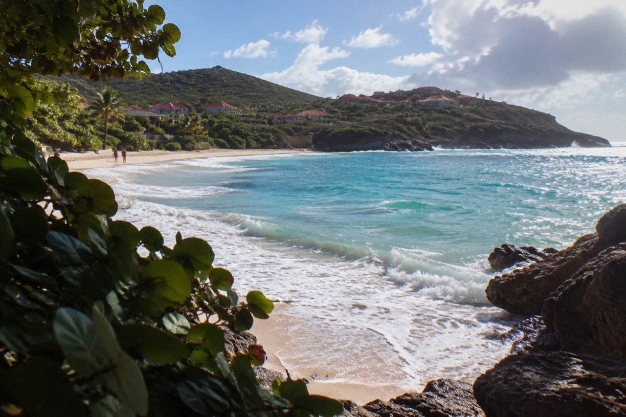 A curve of sand on a Caribbean island shoreline with low green hills behind and small waves reaching the waterline