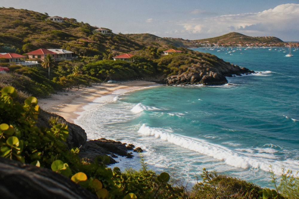 An aerial view of a small Caribbean island coastline with green hills, a sandy cove and turquoise water