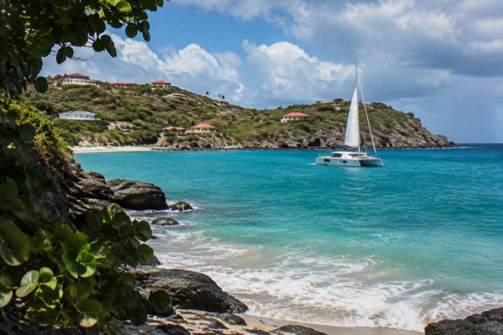 A sailing catamaran on turquoise Caribbean water near a rocky green island headland