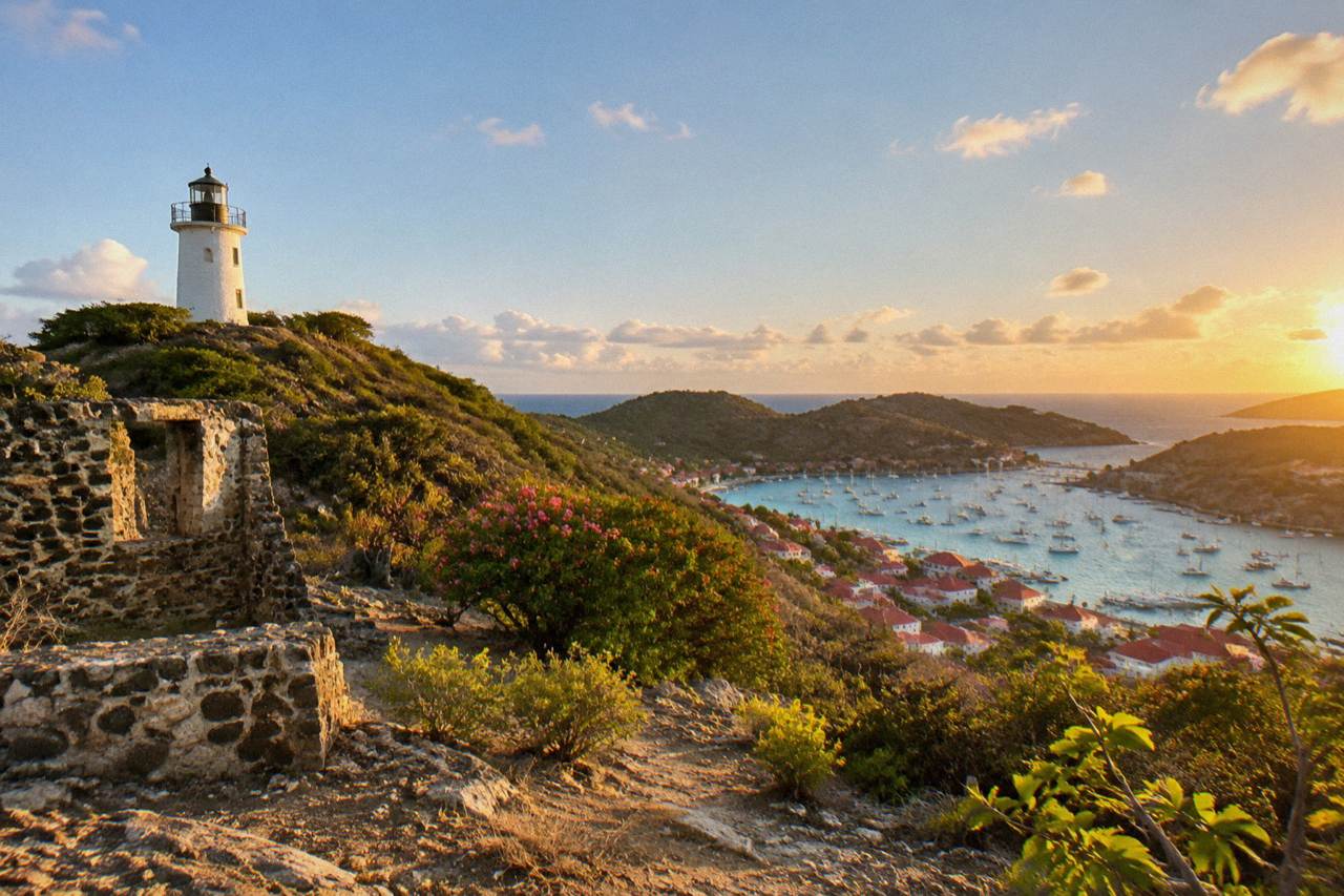 A stone lighthouse tower standing on a green hillside above the red roofs of a Caribbean harbour town