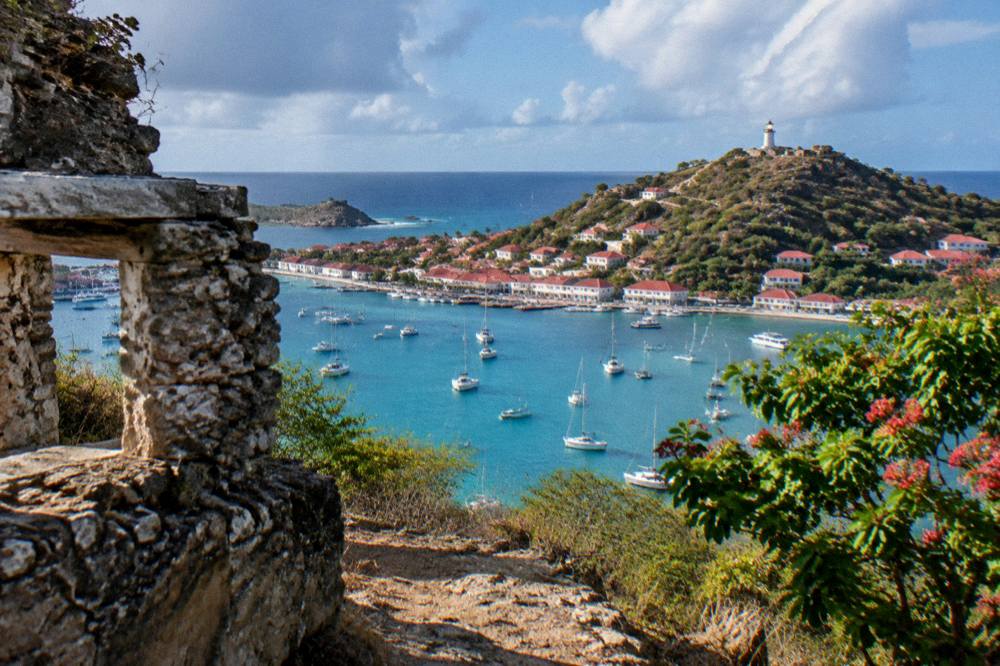 A small Caribbean town at dusk with a lit lighthouse tower on the hillside above tiled roofs