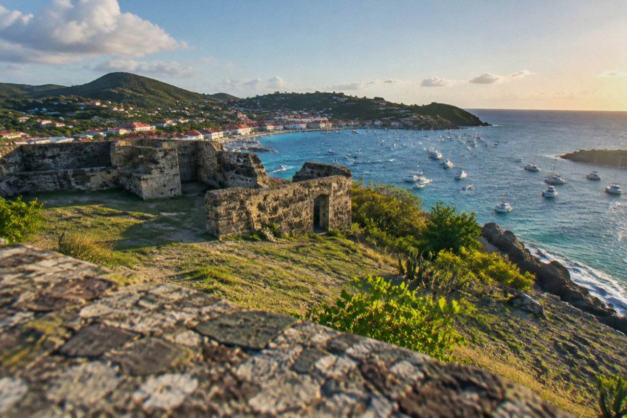 Stone ruins of a small fort on a grassy slope above a rocky Caribbean shoreline and harbor town