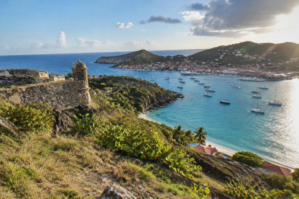Panoramic view over a Caribbean harbor town with red roofs and moored sailboats seen from a grassy headland