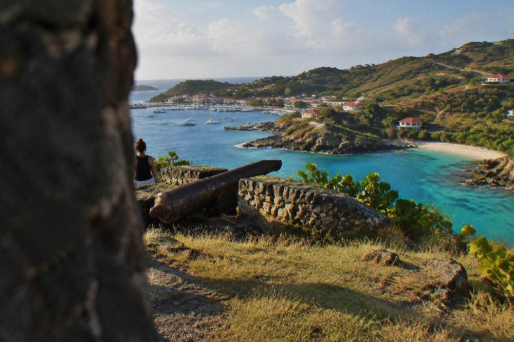 Small rocky beach with clear turquoise water beneath a grassy headland near a harbor town