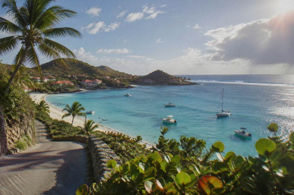 A narrow coastal road with green hills dropping toward a bay with shallow water