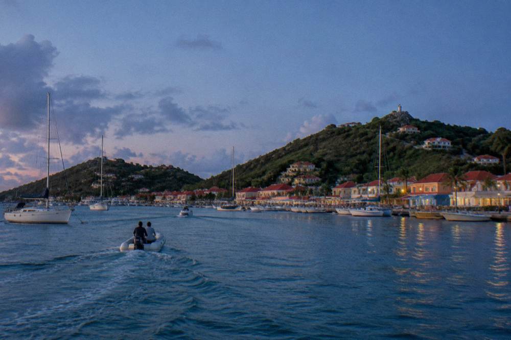 Small tender boats crossing open water toward a harbour town with hills behind