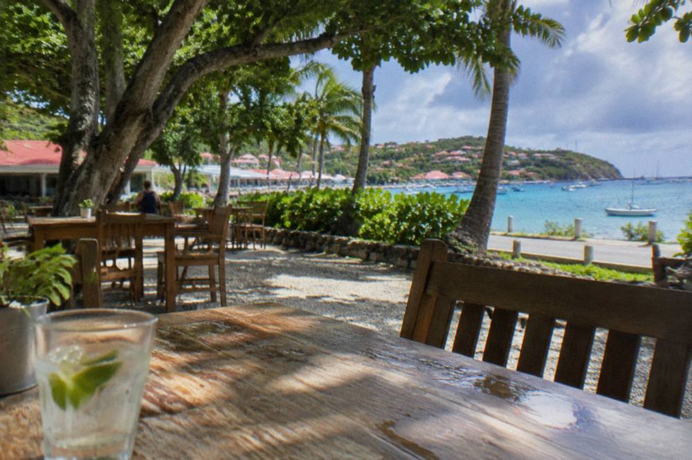 Simple wooden tables and chairs scattered on gravel under large shade trees beside a Caribbean street