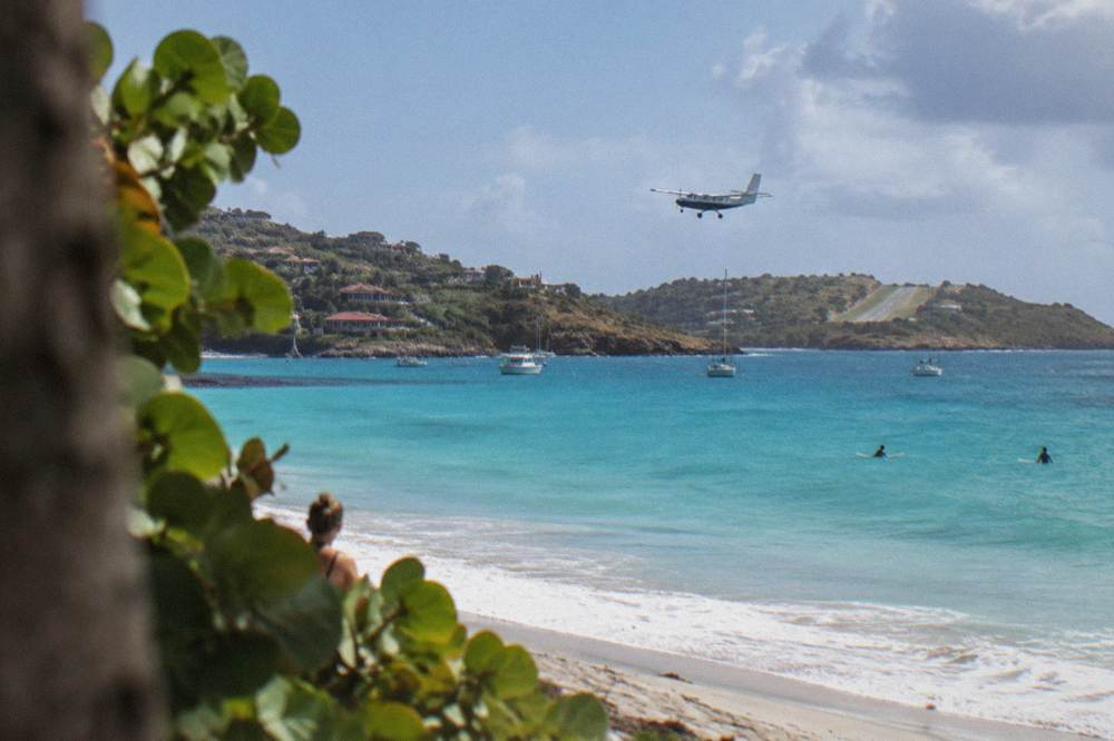 A small single-engine turboprop airplane flying low over a Caribbean bay toward a hillside runway