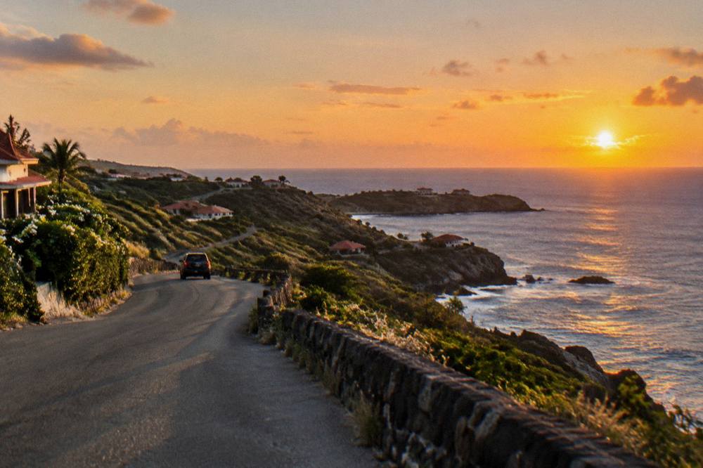 A hillside road at dusk with the sky turning orange over the sea