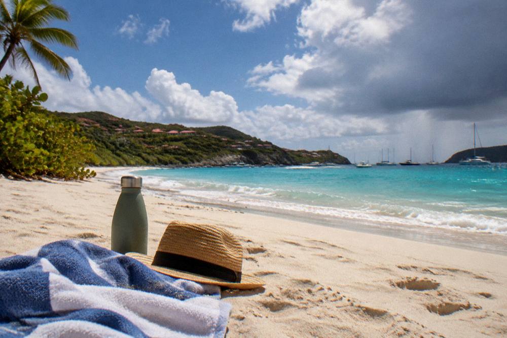 A folded towel, hat and water bottle resting on sand with gentle waves in the background