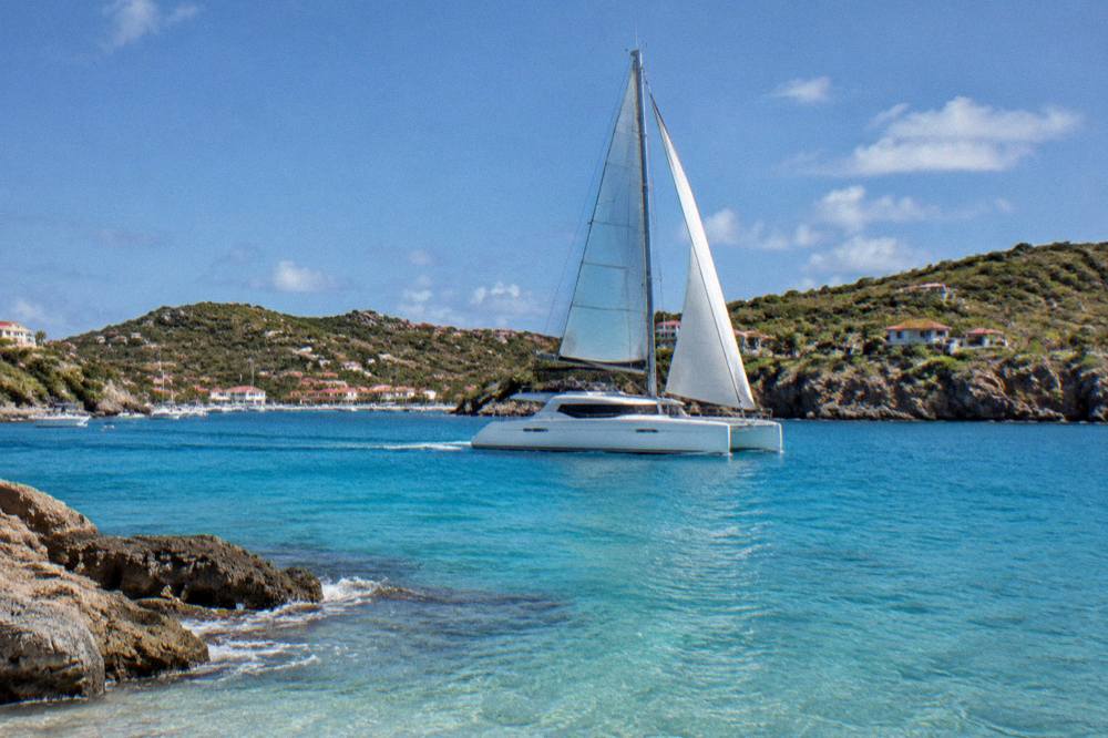 A sailing catamaran cruising along a rocky green coastline in clear blue water