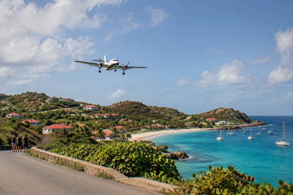 A low-winged turboprop passing over a narrow coastal road with onlookers standing nearby