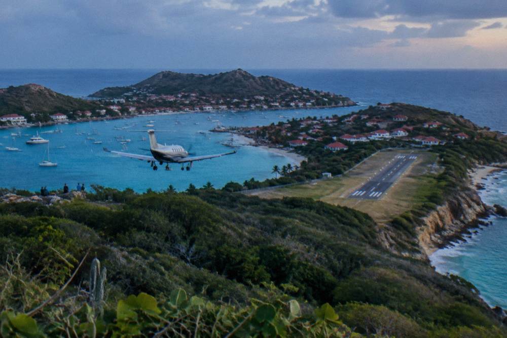 A Pilatus-style single-engine turboprop banking low over green hills toward a coastal airstrip