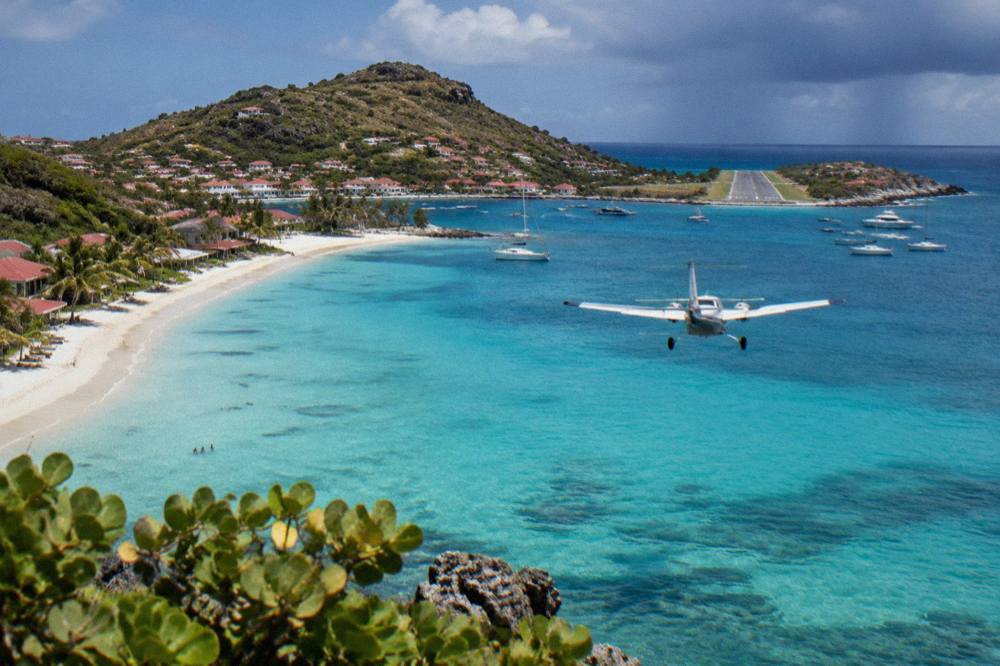 A small twin-engine aircraft flying low over a turquoise bay toward a runway