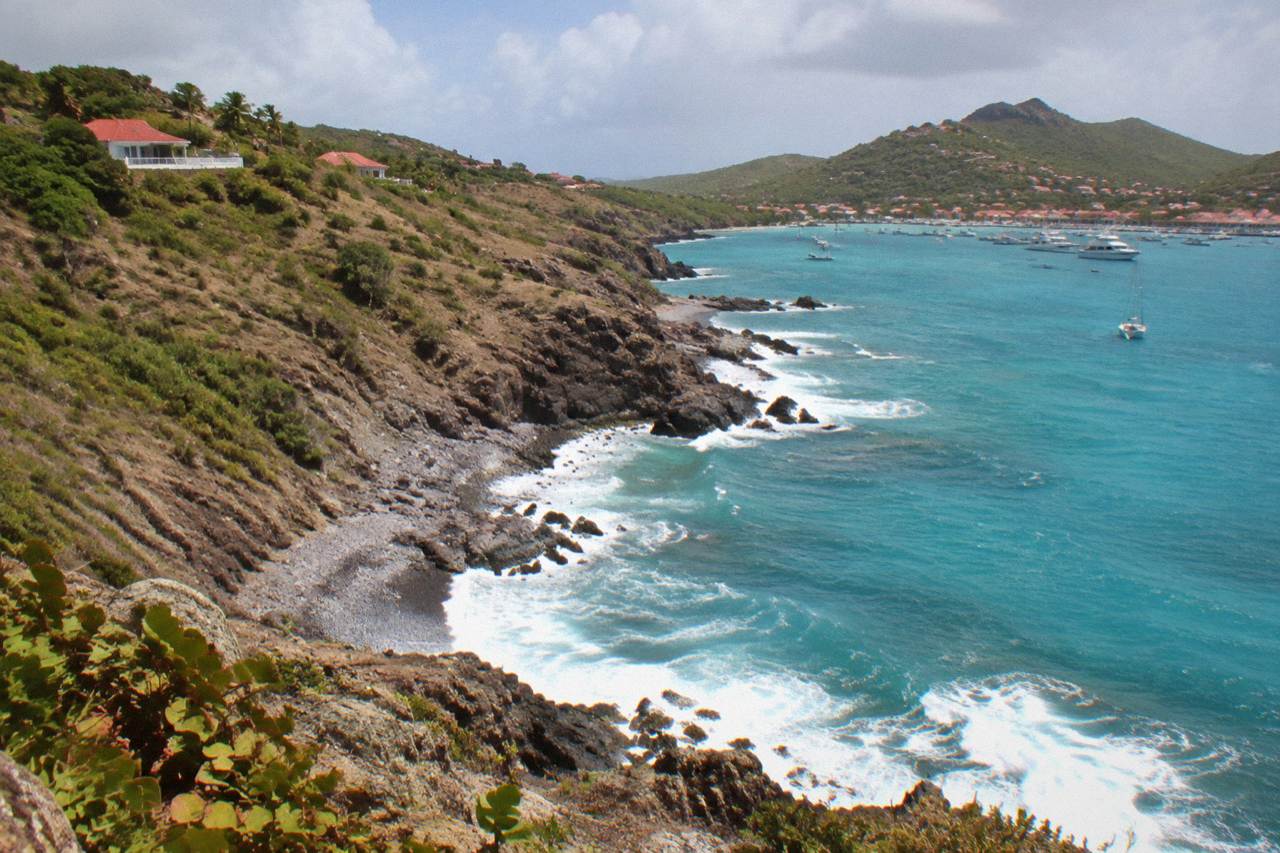 Rocky coastline with green hills dropping toward a turquoise bay, a few white houses on the ridge above