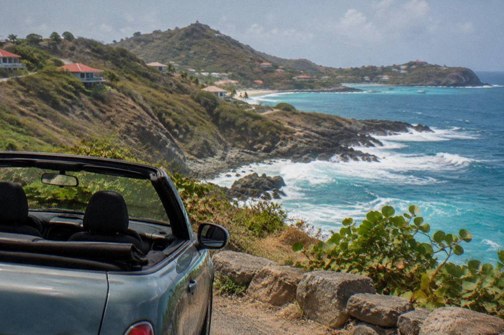 A small open-top rental car parked at a roadside viewpoint above a bay