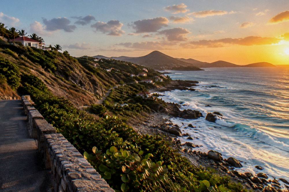 A hillside road viewpoint looking along a coastline toward distant hills at sunset