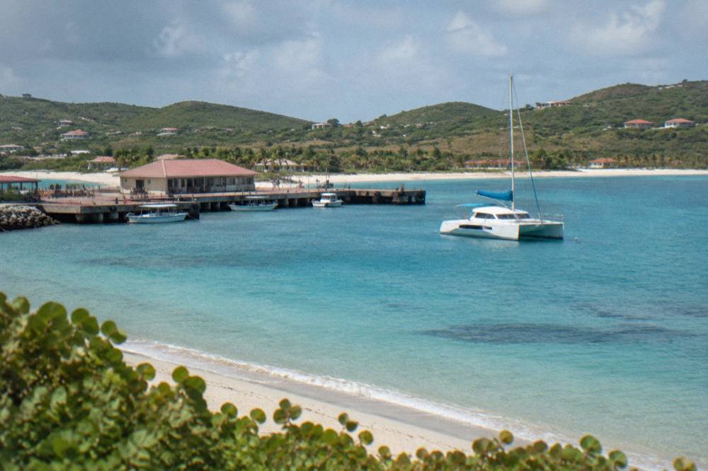 A catamaran moored in a calm bay with a sandy beach and low green hills behind it