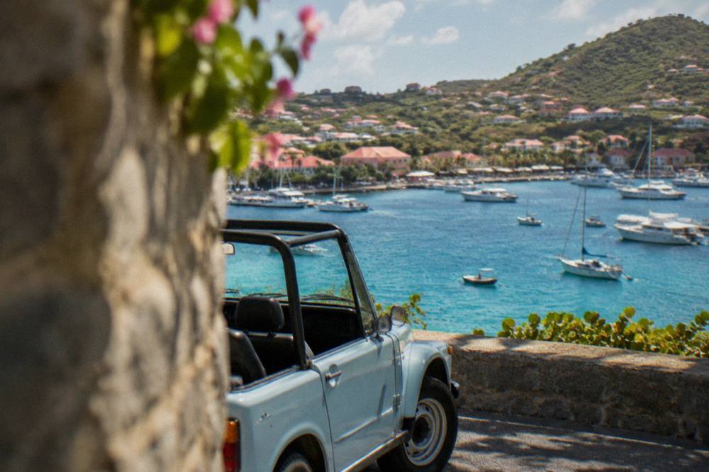 An open-topped small car parked on a hillside road above a turquoise bay