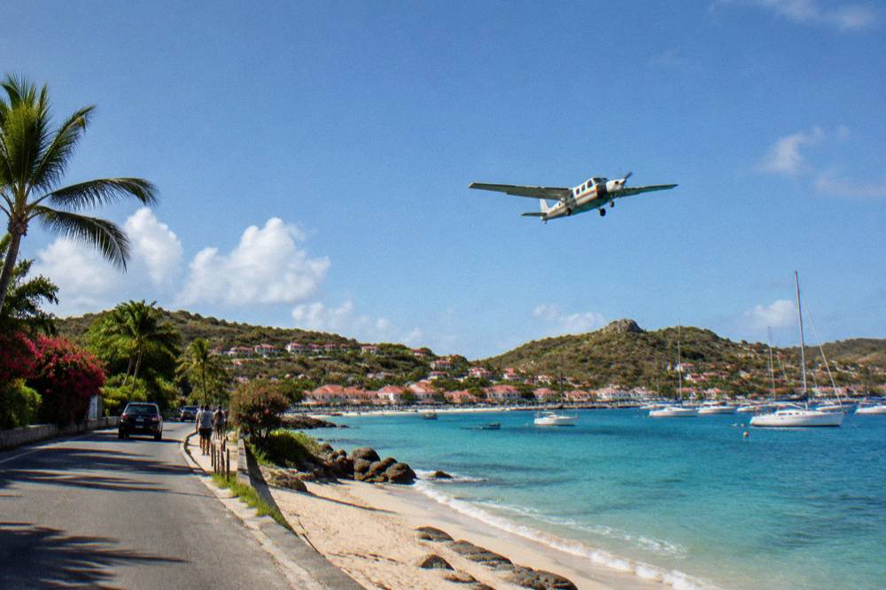 A small turboprop aircraft flying low over a beach road toward a short Caribbean runway