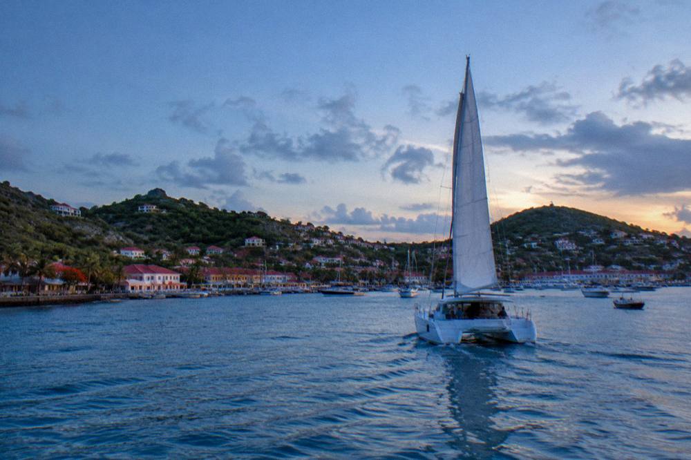 A catamaran under sail approaching a green Caribbean island