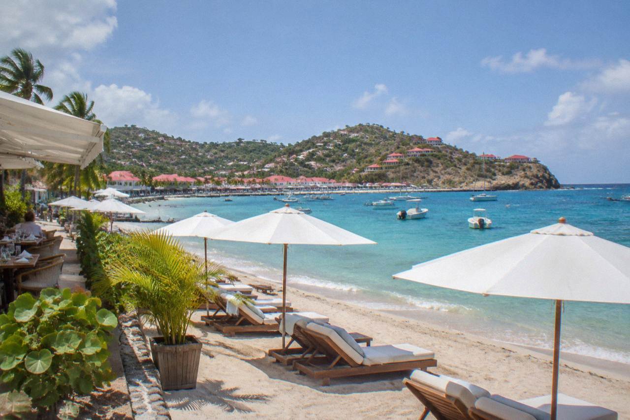 A beach restaurant terrace with white umbrellas and lounge chairs facing turquoise water on a Caribbean island