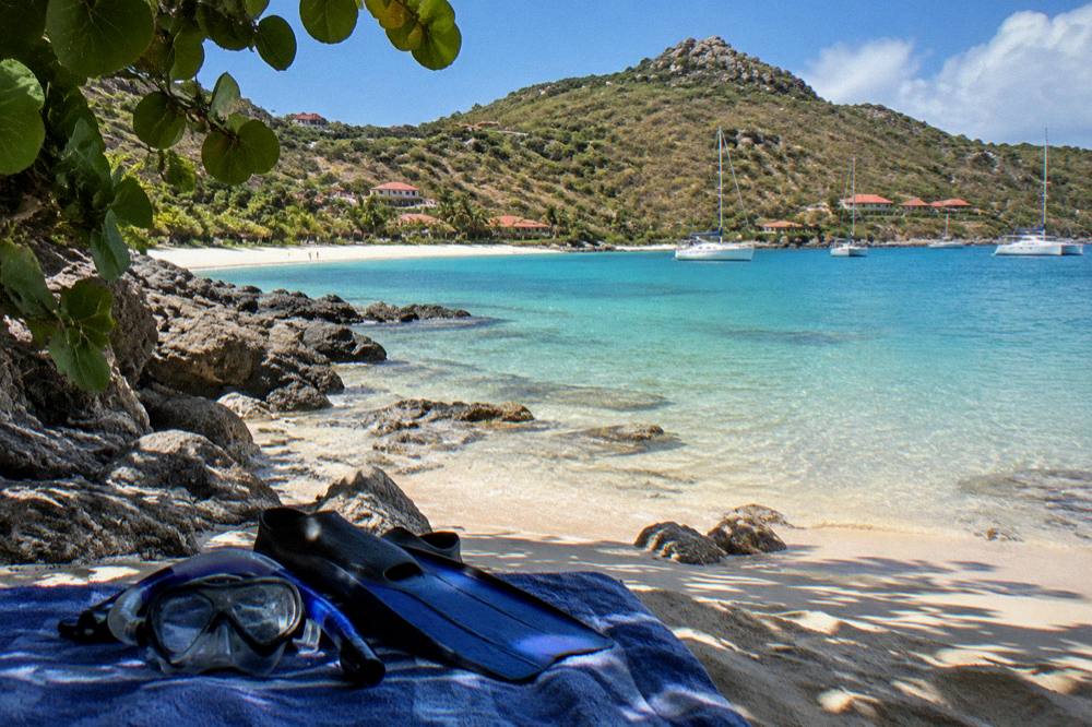 A snorkel mask and fins resting on a towel on sand beside a rocky shoreline