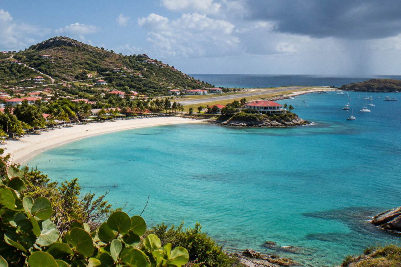 A hotel building on a rocky point above a curved turquoise bay with a landing strip visible along the sand